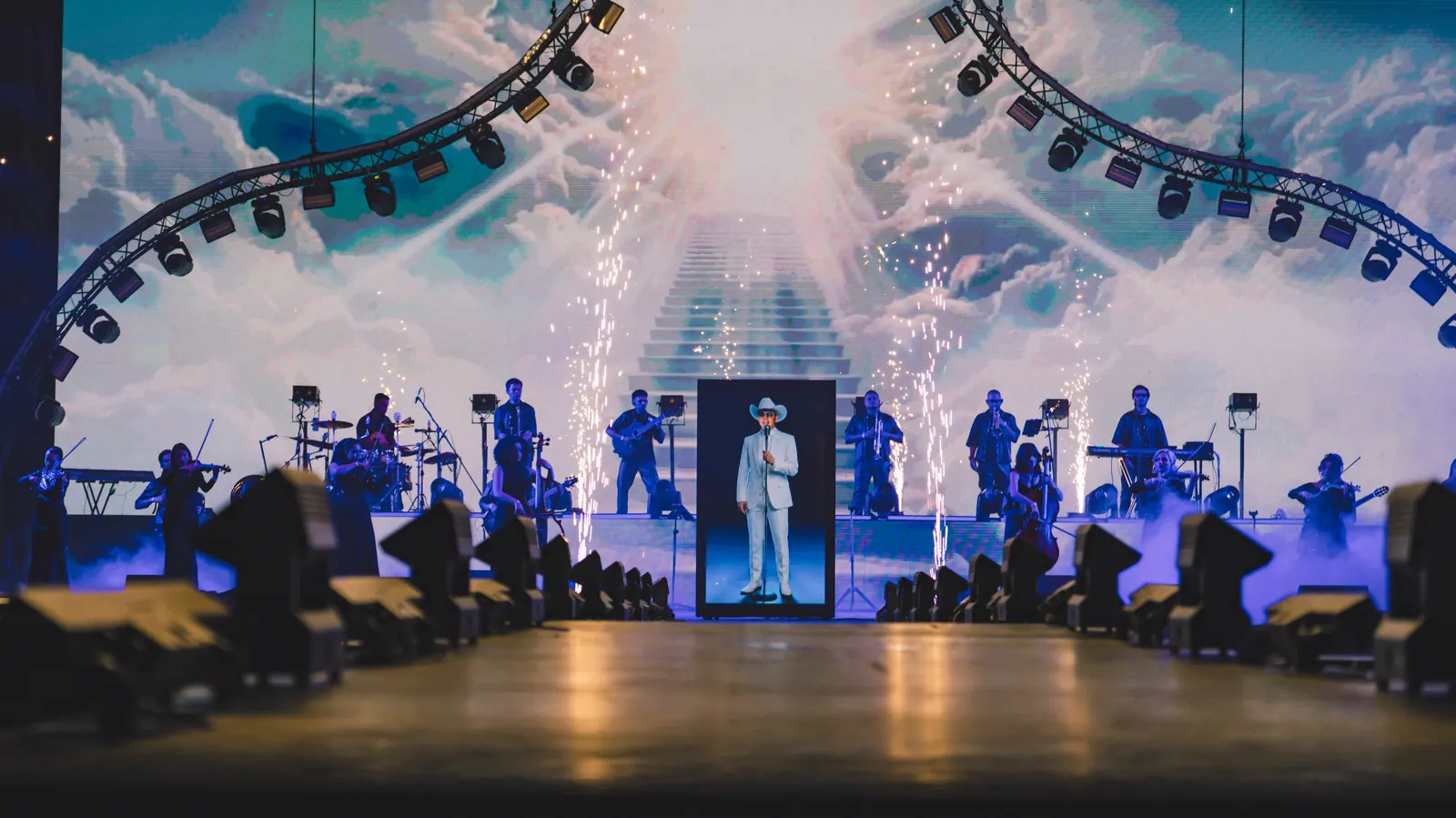 Escenario del tributo en El Campín: banda en vivo, avatar en pantalla vertical y visual de escalera entre nubes